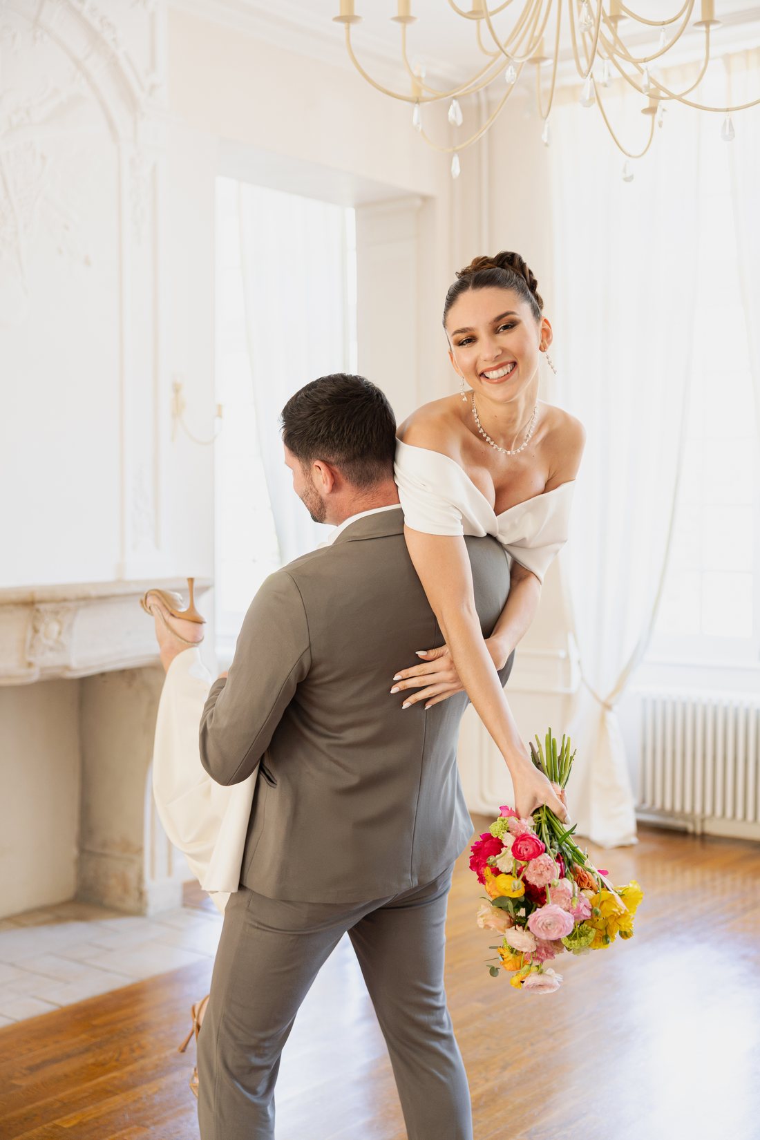 Mariée souriante en robe à encolure bateau, portée par son marié, un bouquet coloré à la main