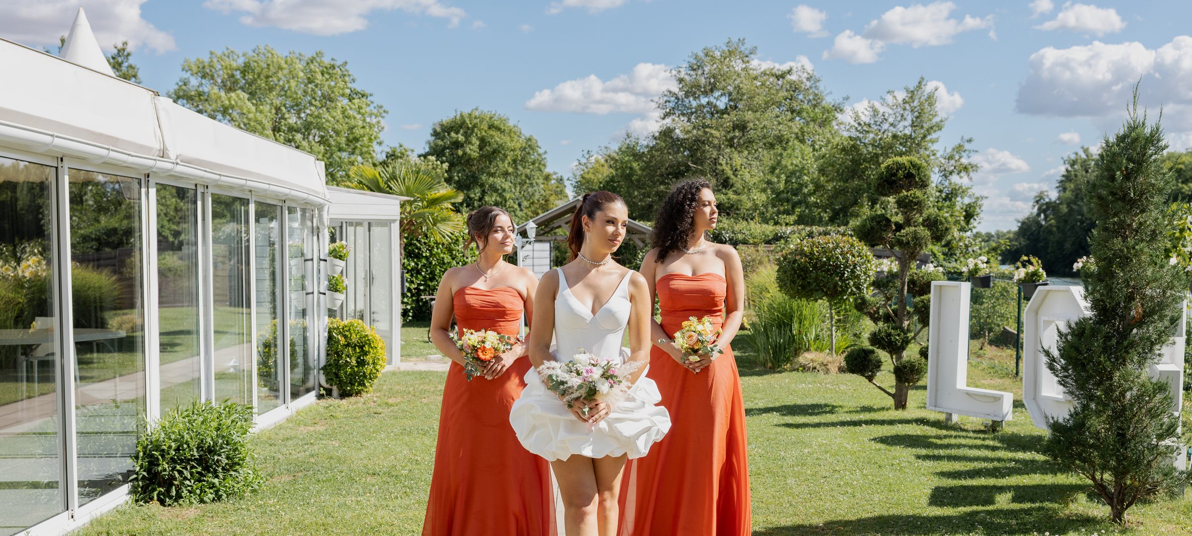 Une mariée entre deux demoiselles d'honneur en robes orange, dans un jardin